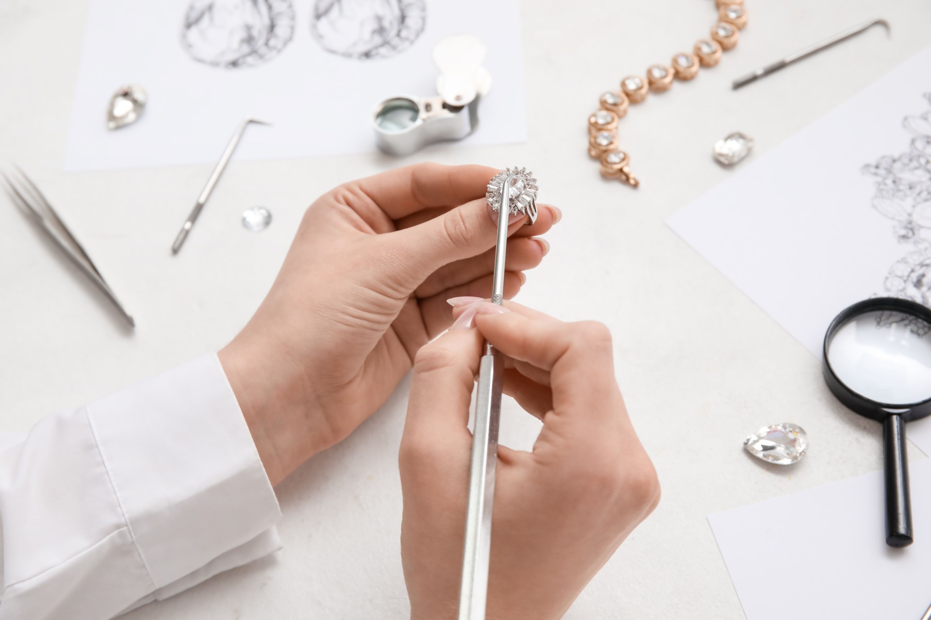 Jeweler's hands meticulously working on a diamond ring with specialized tools and sketches